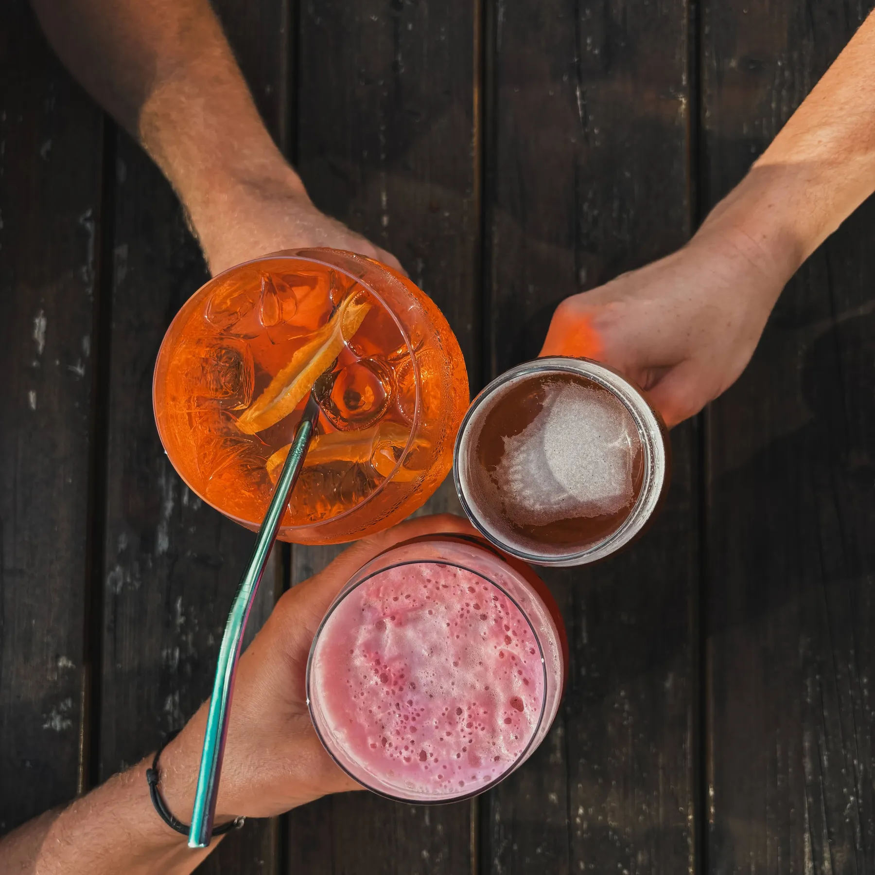 Trois verres levés au-dessus d’une table du Molly’s Pub : un spritz, un cocktail rose et une bière